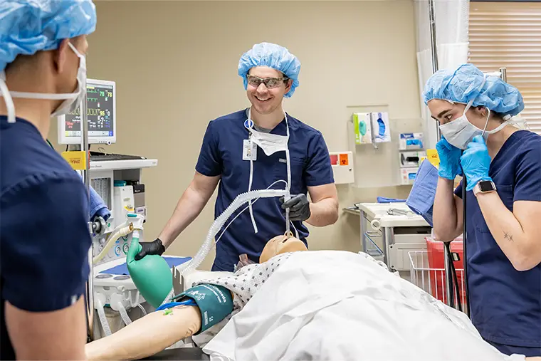 Nick Houge in the Simulation Center with a maniken on the table. Two other students with masks assist.