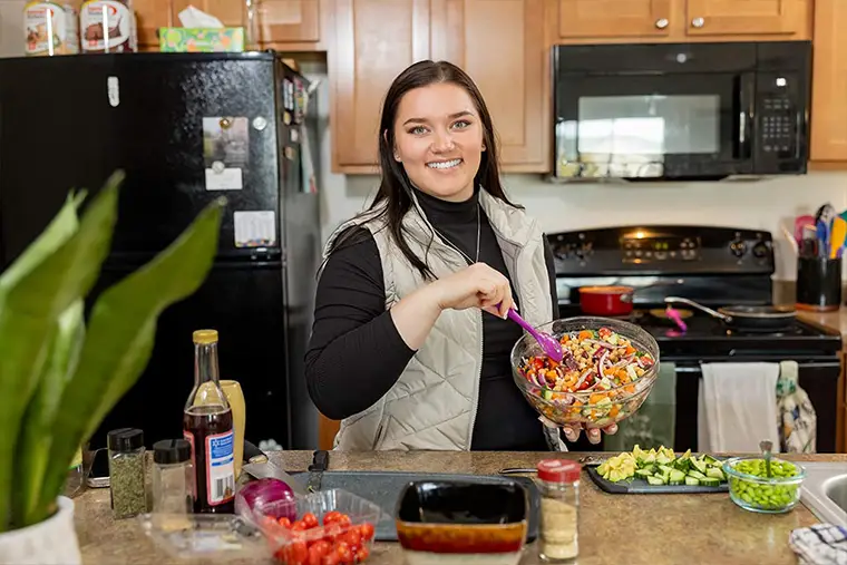 McKenzie Moe smiles while preparing a colorful salad in a home kitchen. She stands at the counter holding a glass bowl and mixing with a purple spoon. Fresh ingredients like cherry tomatoes, chopped cucumbers, and edamame are spread across the counter. The kitchen features light wood cabinets, a black refrigerator, and a black stove and microwave. A green houseplant is visible in the foreground.