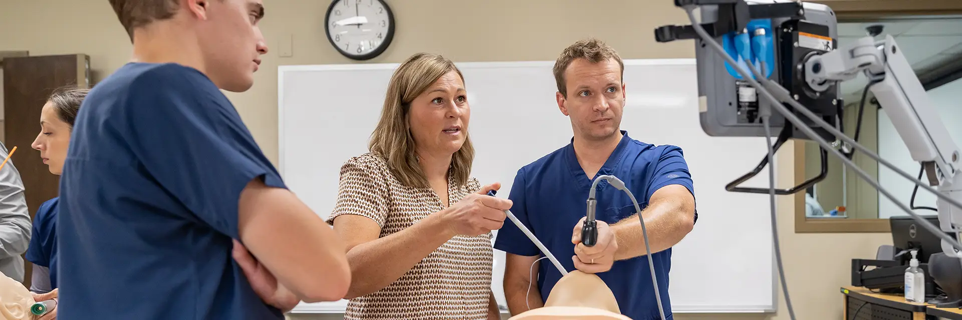two students in blue scrubs stand before a monitor as the student on the right inserts a breathing device. An instructor looks on, holding a tube.