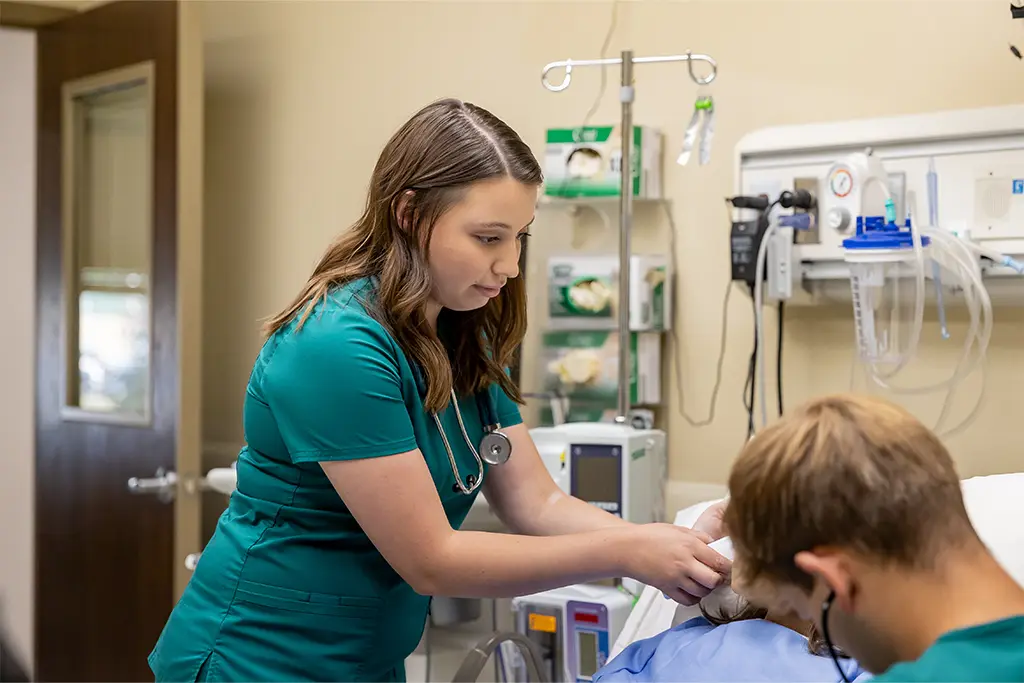 A nursing student in green scrubs practices a medical procedure on a patient simulator in a clinical setting, focused on hands-on learning. Medical equipment and monitoring devices are visible in the background.