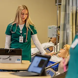A nursing student stands at the bedside observing a maniken simulator