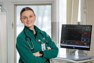 Jenna in green scrubs in a hospital room. A patient monitor blips off to the side