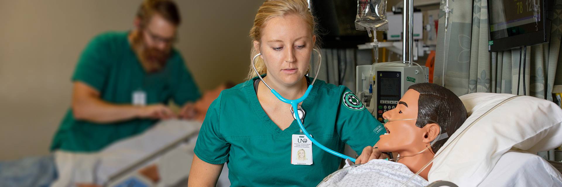 a student in green UND scrubs uses a stethoscope to check the heart rate on a maniken. The maniken is lieing on a hospital bed. Another student works on the same task with a maniken in the background.