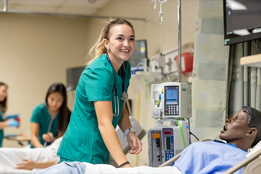 A nursing student in green scrubs leans over the bed of a simulation maniken as students work in the background
