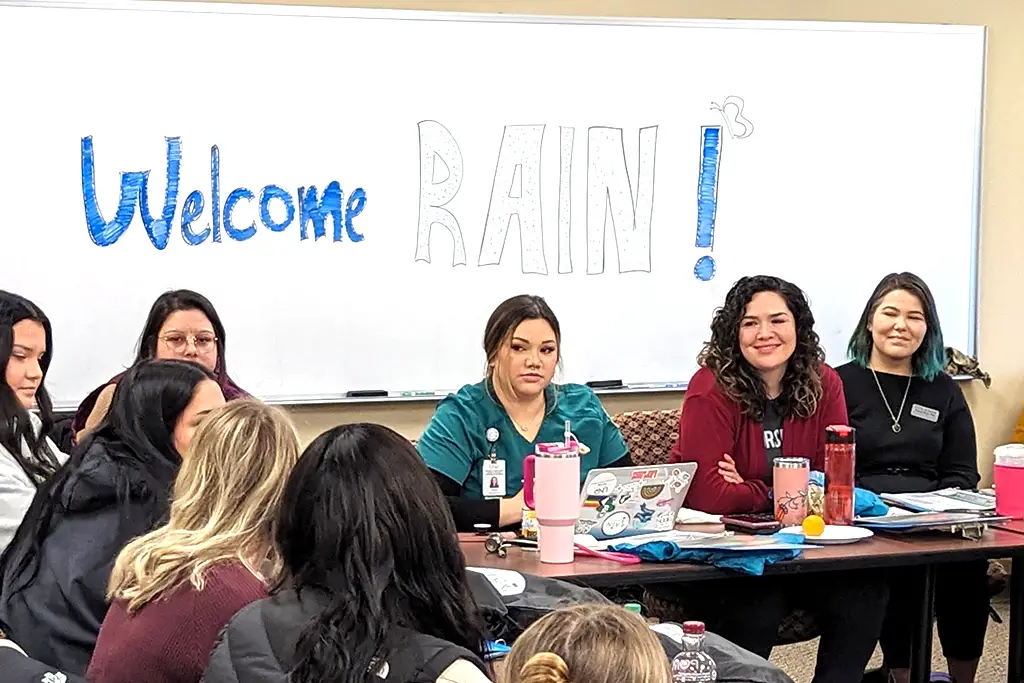 Students and mentors sit around a square table during an orientation event. the words "Welcome to RAIN" are on the board behind them