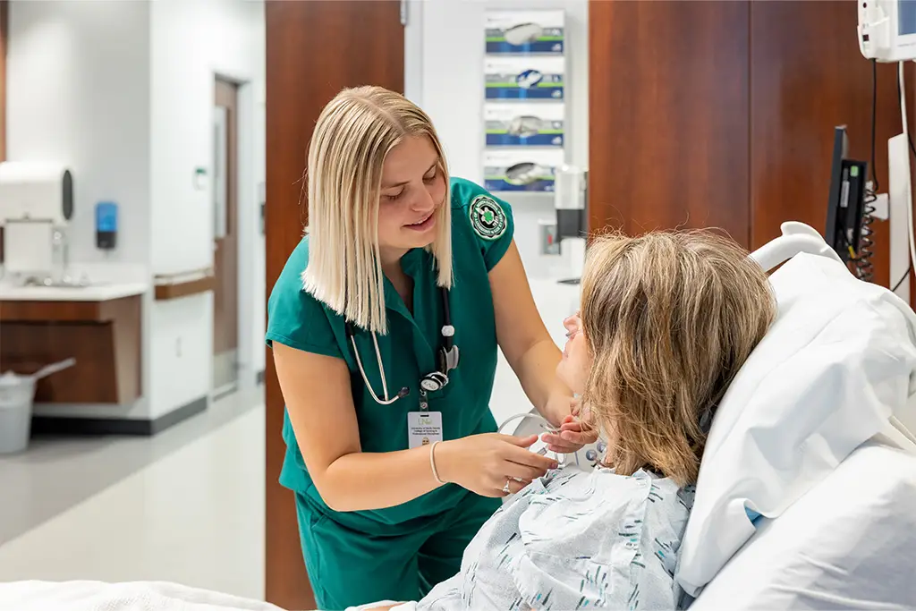 a nursing student in green scrubs adjusts a breathing tube on a patient who is lying in a hospital bed