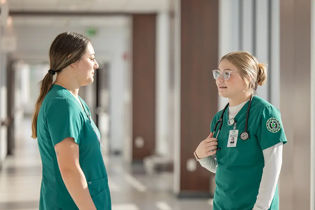 Two nursing students in green scrubs pause to talk in a hallway